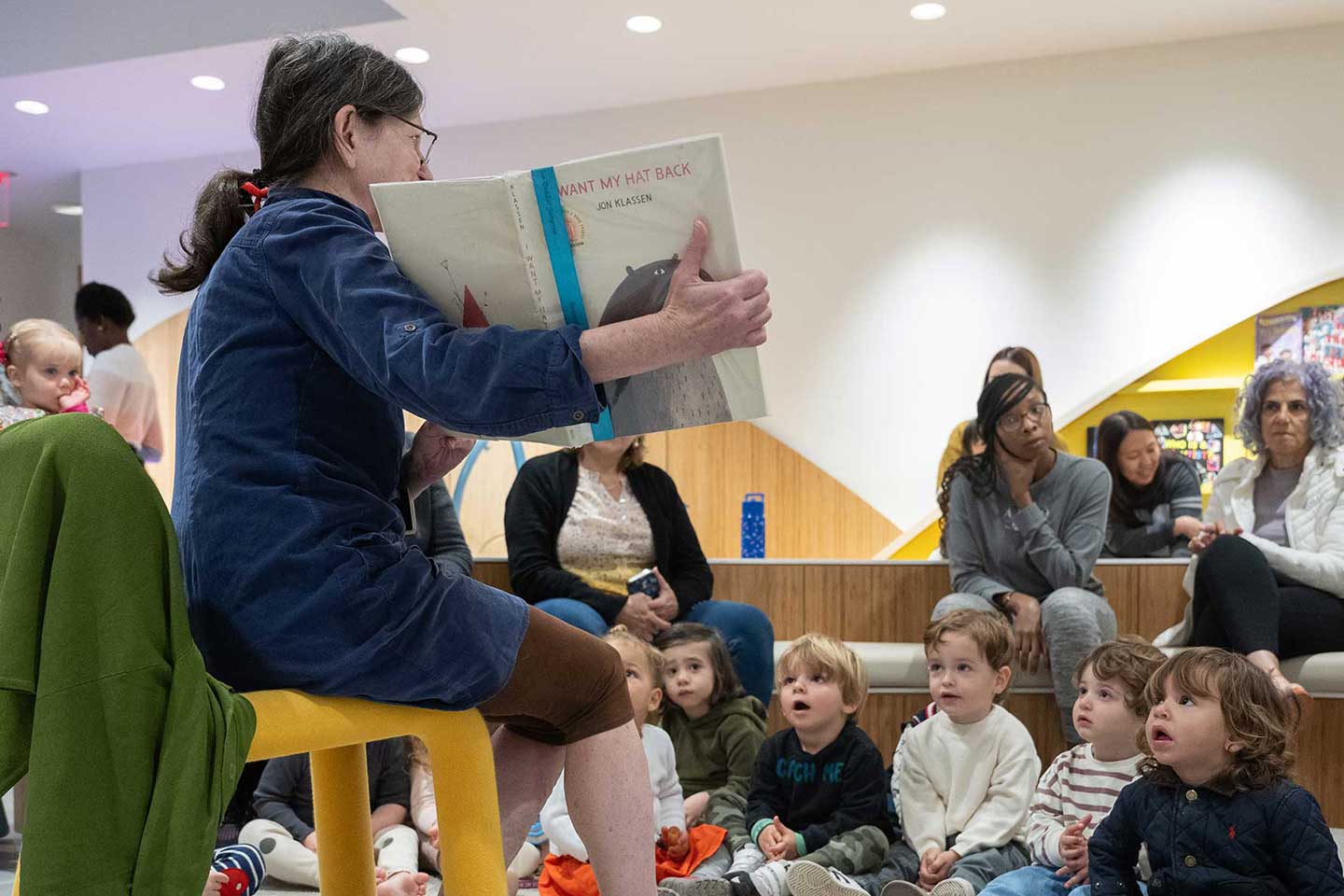 A woman sits on a yellow chair. She is holding open a picture book, and a group of toddlers seated before her on the floor are listening to her reading the story.